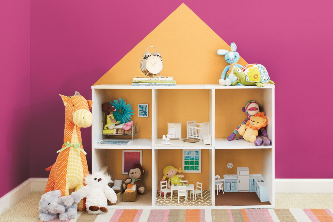 A dollhouse with stuffed animals sits in front of a deep pink wall in a lived-in kids’ bedroom. 
