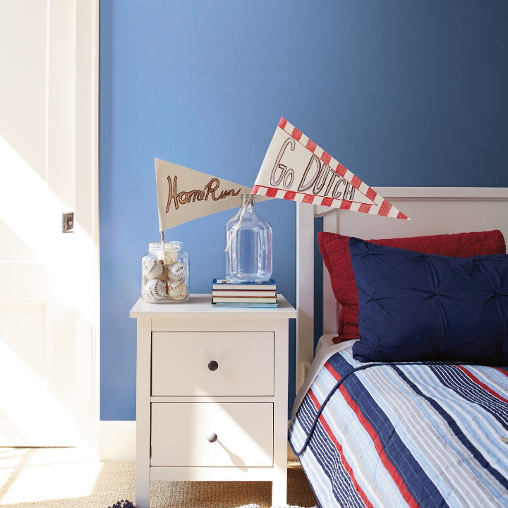 Colorful children’s bedroom with team pennants in vases on a side table with books. Walls painted blue jay blue.