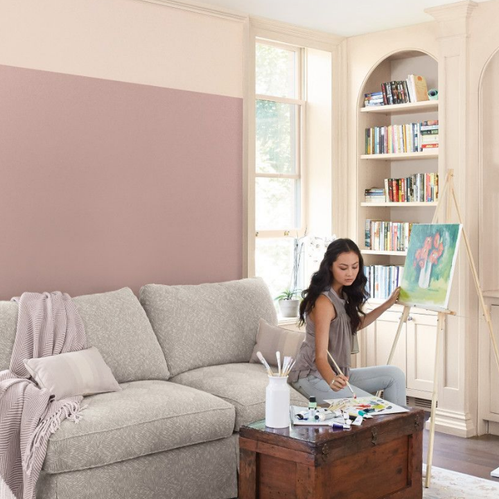 A woman sits on a couch, painting on an easel in a living room, walls painted neutral hennepin stone.