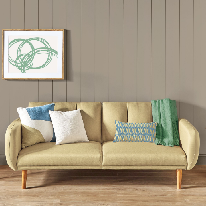 Yellow couch on a light wood floor sits in front of a neutral rustic greige wall with framed white and green abstract art.
