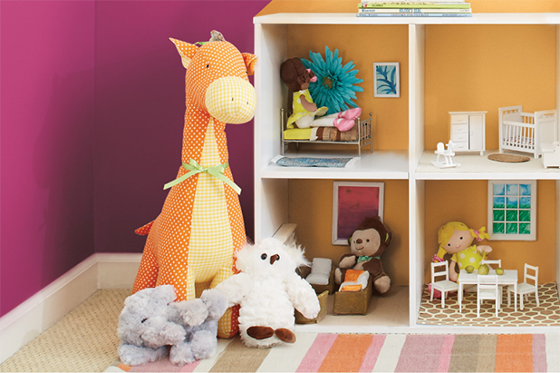 A close shot of a stuffed giraffe, bird and elephant sit adjacent to a dollhouse with other dolls inside.