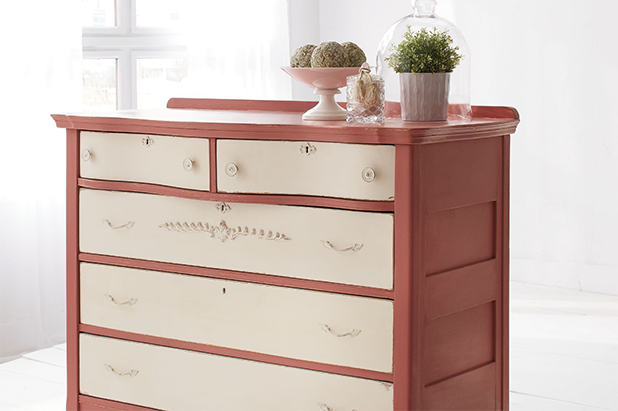 A dresser sits in a white room. Its drawers are painted a chalky beige and the dresser itself is painted a chalky red.