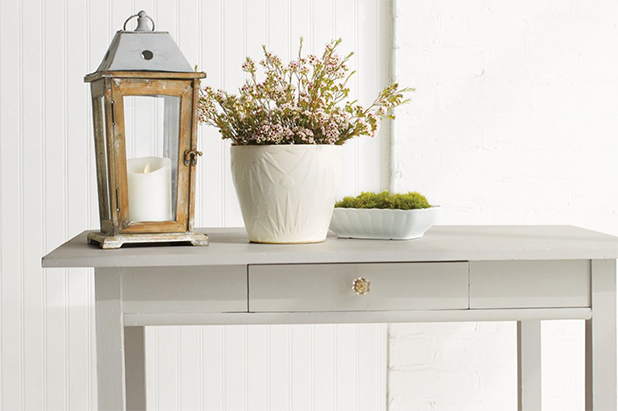 A chalky white console table with a candle lantern, potted plant and potted moss sits in front of a white wall.