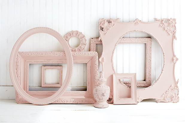 A variety of different shaped empty art frames, painted chalky pink, sit on a white floor resting against a white wall.