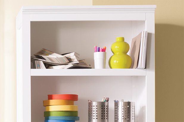 A white cabinet against an off-white and beige wall. Paint chips, pen holders, vases and rolls of tape on shelves.