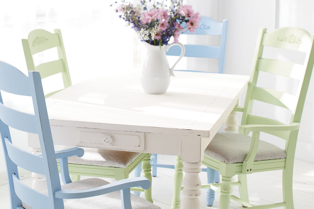 A white square dining table with a vase holding wildflowers sits in a sunlit room, with light green and blue chairs around.