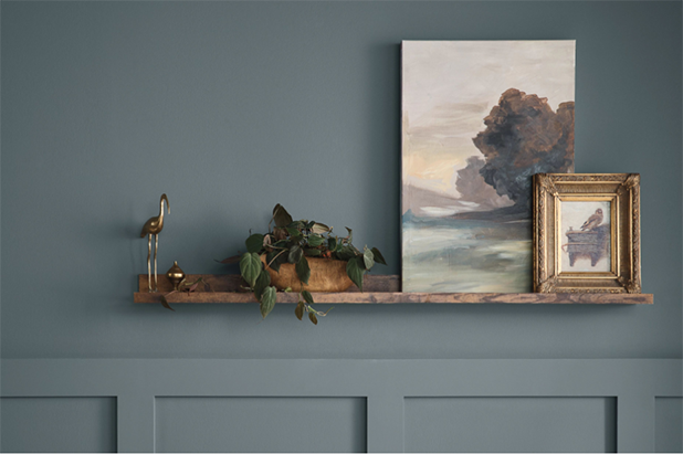 Deep blue wall with wainscoting and a dark wood shelf with paintings and a plant sitting on top.