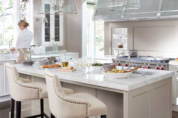Woman washes dishes in large kitchen with island, pendant lights and a backsplash painted sultry gray.