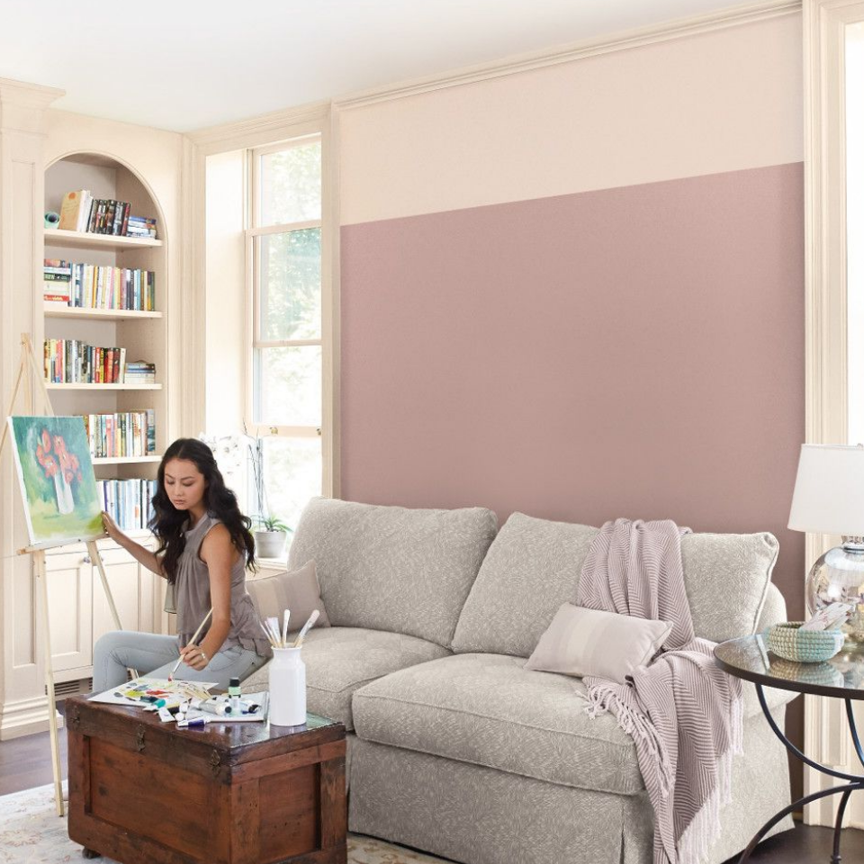 A young woman sits on a couch, painting on an easel in a living room, walls painted the color neutral hennepin stone.