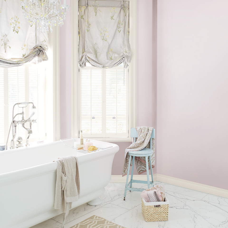 Open, airy bathroom with white claw-foot tub, blue stool with curtains draped over windows. Walls painted misted mauve.