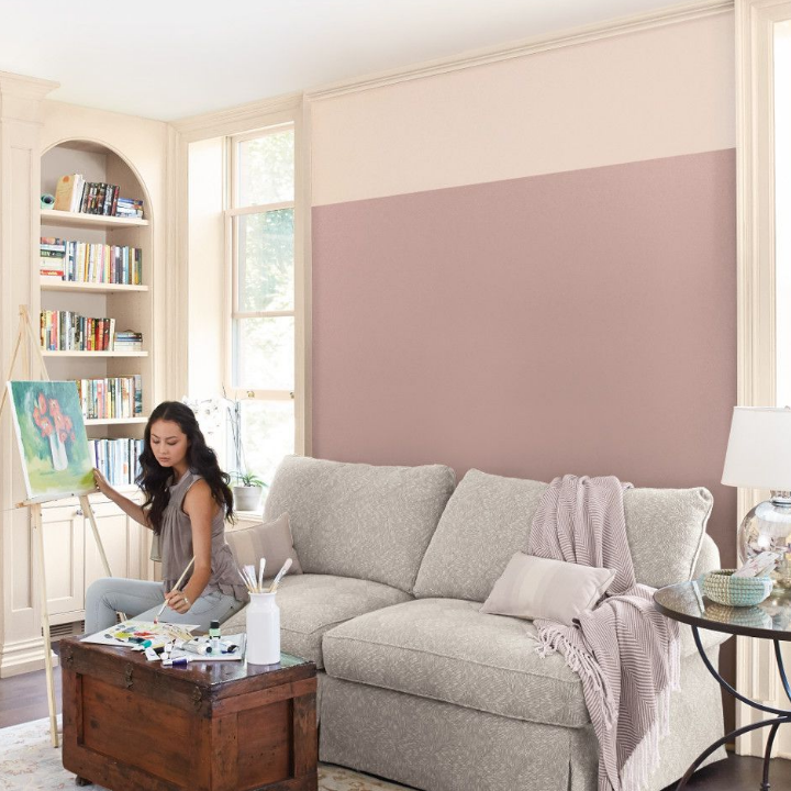 A young woman sits on a couch, painting on an easel in a living room, walls painted the color antiqued fuchsia.